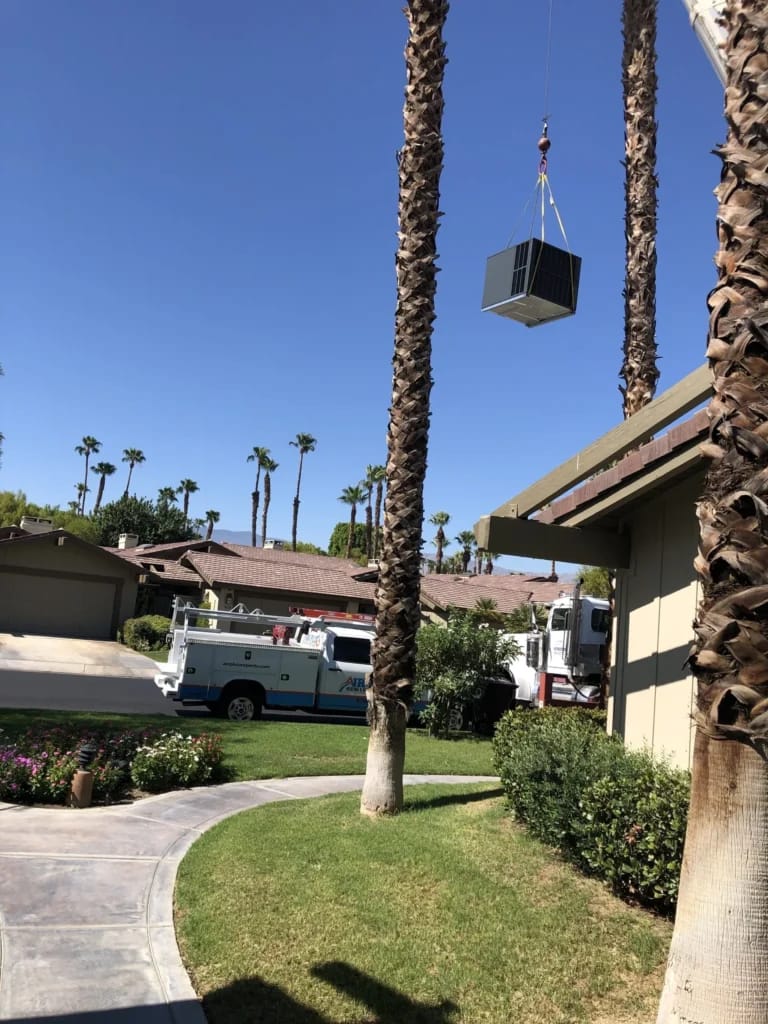 Crane lifting a new residential rooftop HVAC unit over palm trees during an Air Plus heating and air conditioning installation.